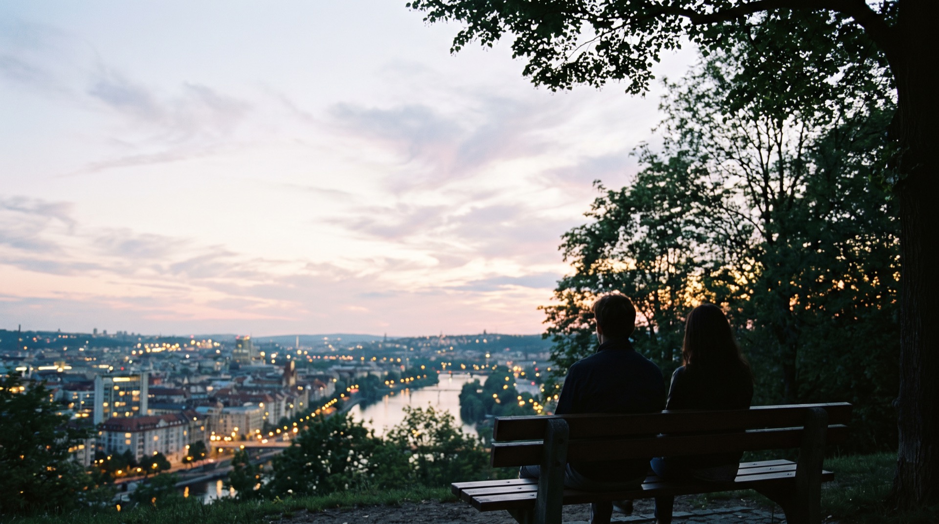 Two people on a park bench at dusk, seen from behind, looking out over a calm river city as its lights come on.